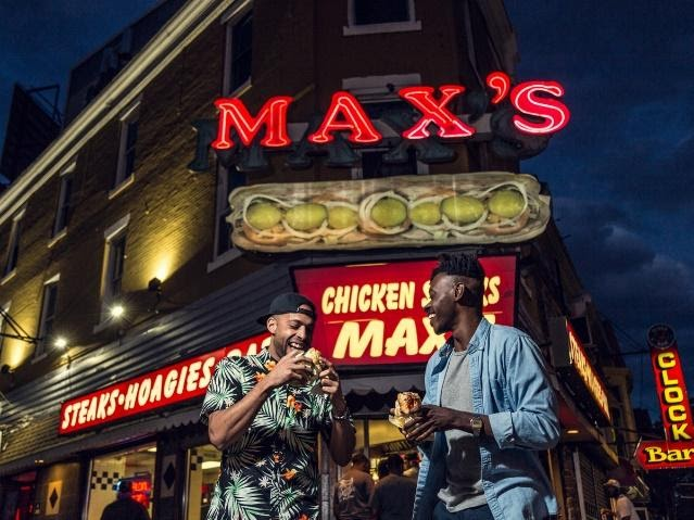 Two people stand in front of Max's Steaks in Philadelphia eating steak sandwiches.