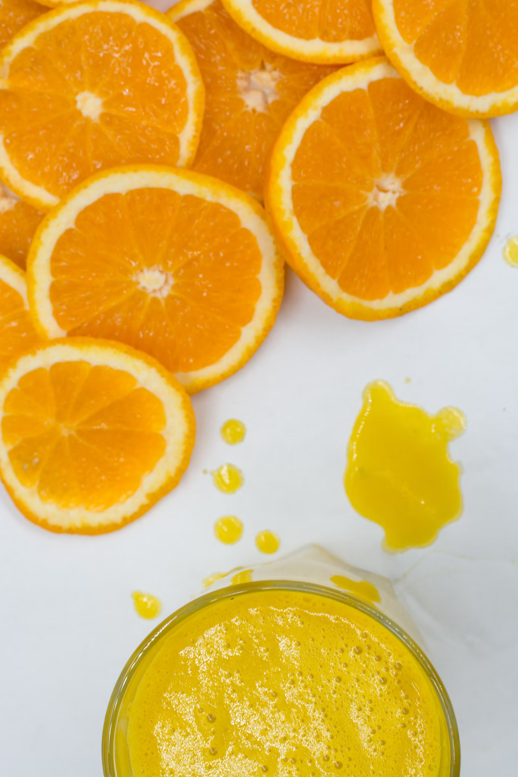 A top-down view of orange slices on a white surface above a cup of fresh orange juice.