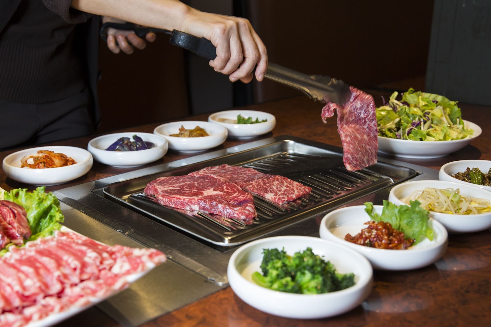 A person holding a cut of beef above a Korean BBQ while two other cuts of beef are cooking. The BBQ is surrounded by various side dishes as well as additional cuts of pork.