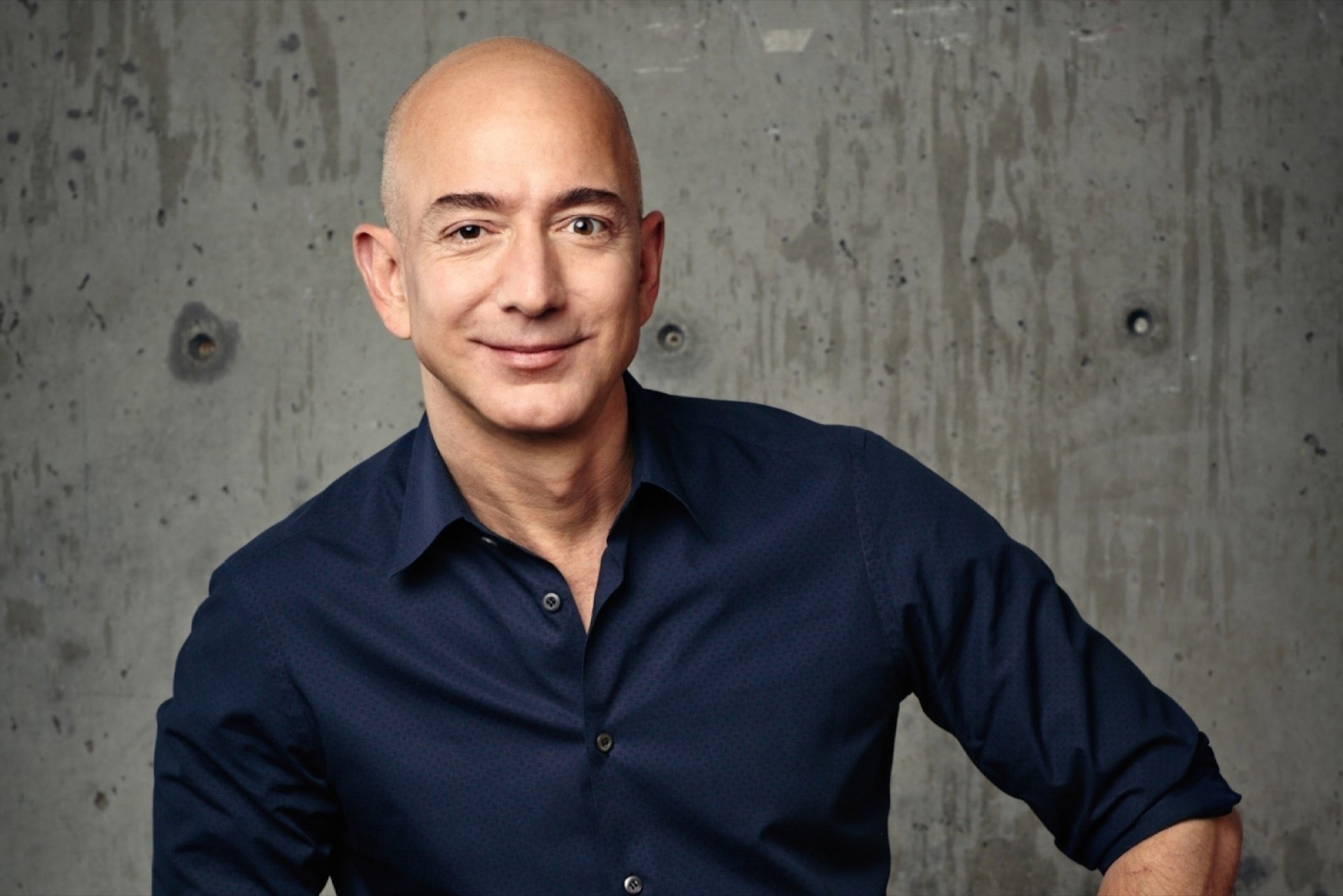 Jeff Bezos, wearing a dark shirt, sits against a concrete background.