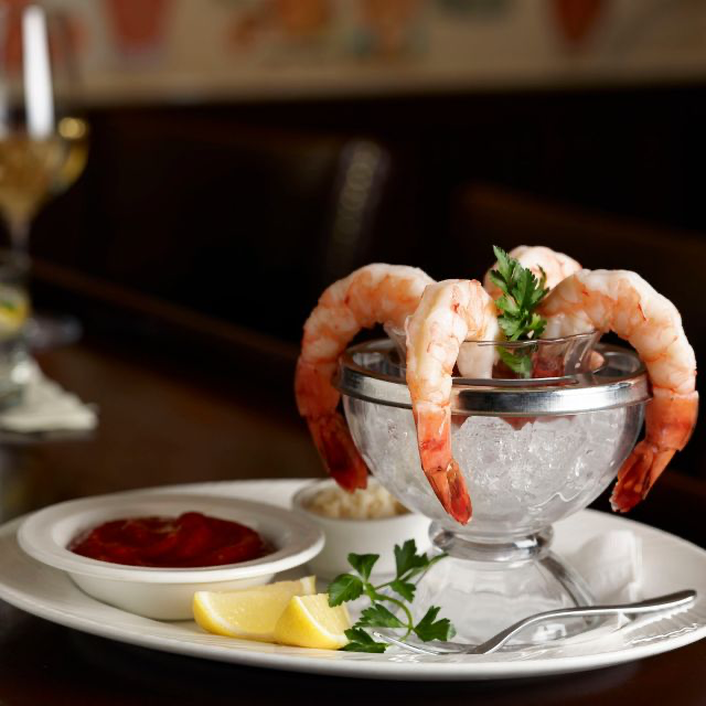 Four shrimp in a serving dish with ice, with a fork, lemon slices, and other side dishes.