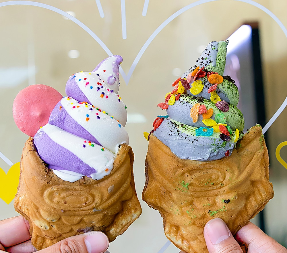 A person holding two ice cream cones filled with multicolored soft serve. The ice cream cones are shaped like fish.