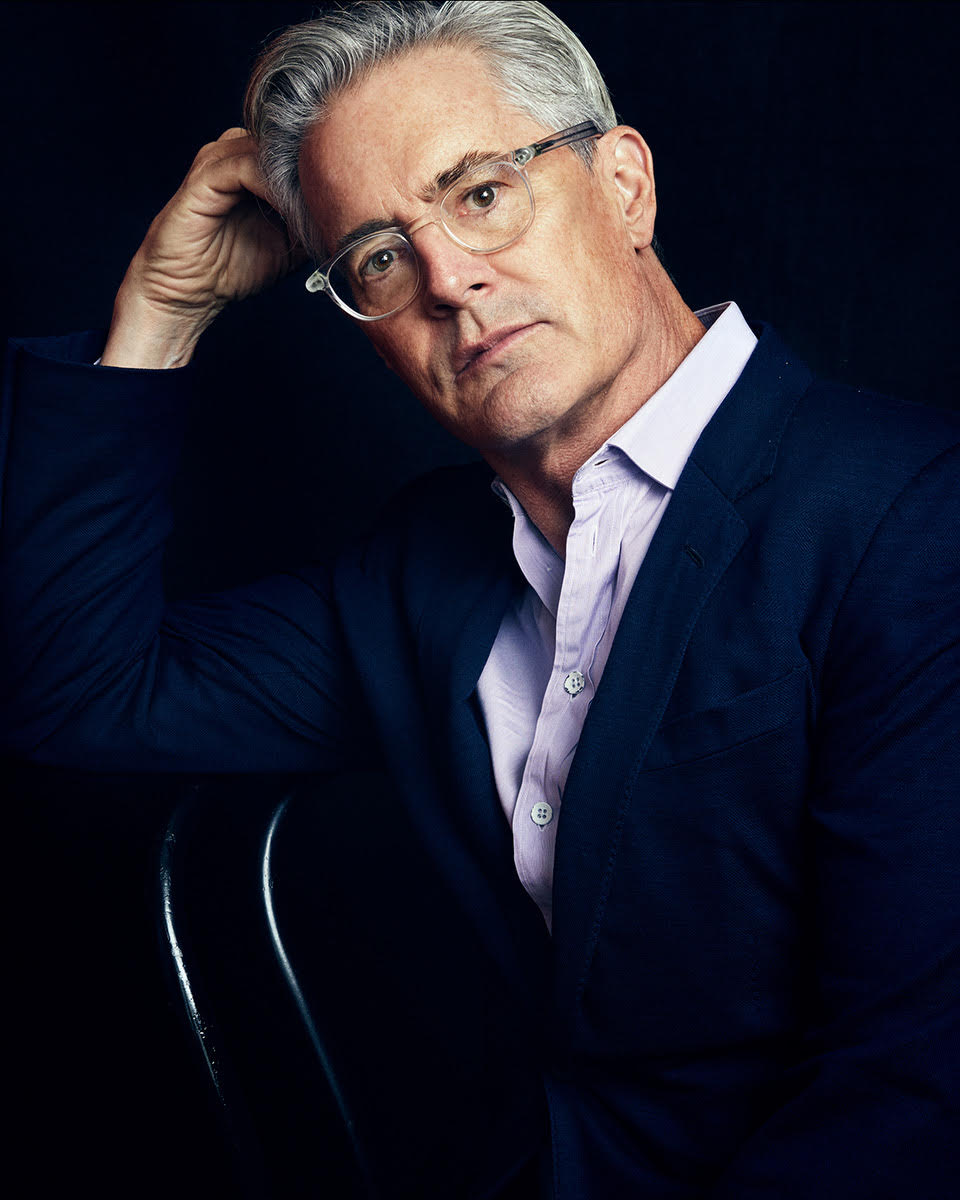 Kyle MacLachlan, wearing a blue blazer and white shirt, leans on a chair back.