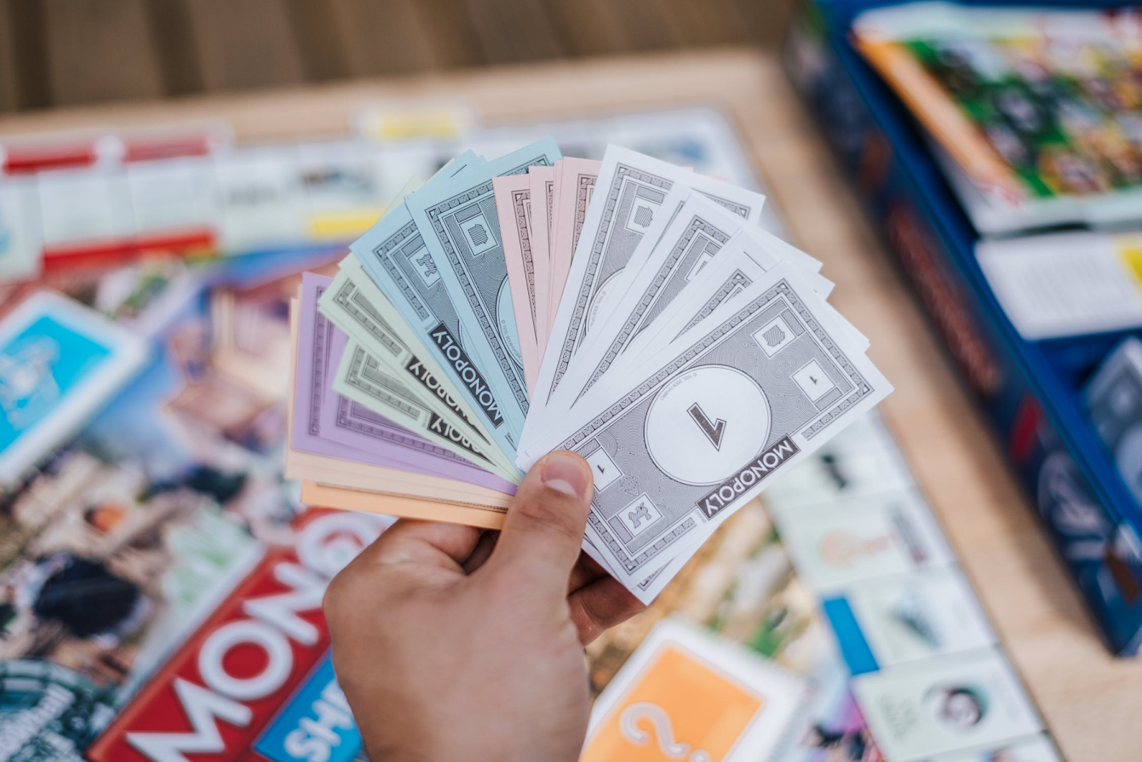 A person holding a bunch of Monopoly notes flared out between their thumb and their fingers with the board game in the background.