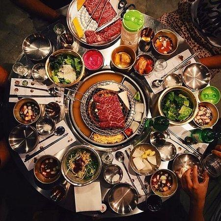 A top-down view of a table laden with different dishes for a Korean BBQ. There is a tabletop BBQ in the center with a cut of meat on it.